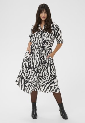 Donna che indossa un abito midi con stampa astratta in bianco e nero con cintura, calze nere e stivaletti neri, in piedi con le mani nelle tasche.