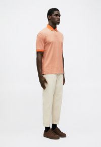 tricou polo cu mânecă scurtă în culoarea coral-portocalie, cu un model geometric, guler contrastant. Asortat cu pantaloni de culoare crem și teniși maronii.