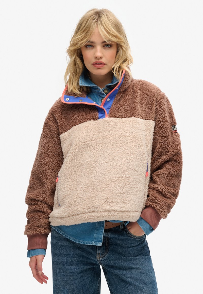 Pull en polaire marron et crème avec un col haut, des accents bleus et des poches latérales. Il présente une texture douce et une fermeture à pression réglable.