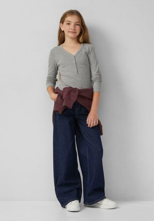 Meisje staat, draagt een grijze lange mouw henley, donkere wijd uitgesneden jeans, witte sneakers en een kastanjebruine sweatshirt om haar taille geknoopt.