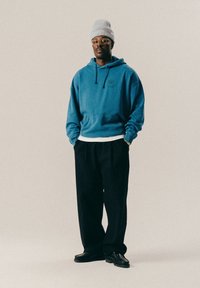 Mężczyzna w szarej czapce, okrągłych okularach, niebieskiej bluzie z kapturem, czarnych szerokich spodniach i czarnych butach stoi z rękami w kieszeniach na jednolitym tle.