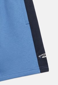 Μπλε και ναυτικό πάνελ υφάσματος με ορατές ραφές και μερικό λευκό λογότυπο Quiksilver κοντά στη ραφή στην ναυτική κορυφή.