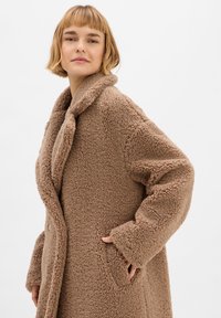 Brauner, fusseliger Fleece-Mantel mit abgerundetem Kragen, langen Ärmeln und Seitentaschen, ausgestattet mit einer weichen, strukturierten Oberfläche und Knopfverschluss.