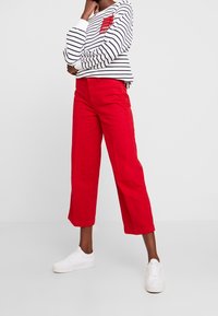 Persona con sneakers bianchi, pantaloni cropped larghi rossi e una maglietta a maniche lunghe bianca e nera a righe con un logo rosso.