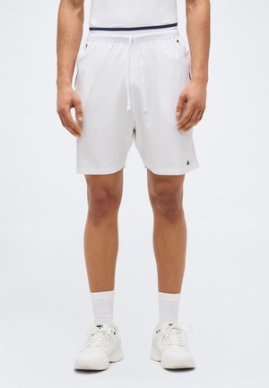 Moški v belih športnih kratkih hlačah s stranskimi žepi, belih nogavicah in belih supergah, stoji ob preprostem ozadju.