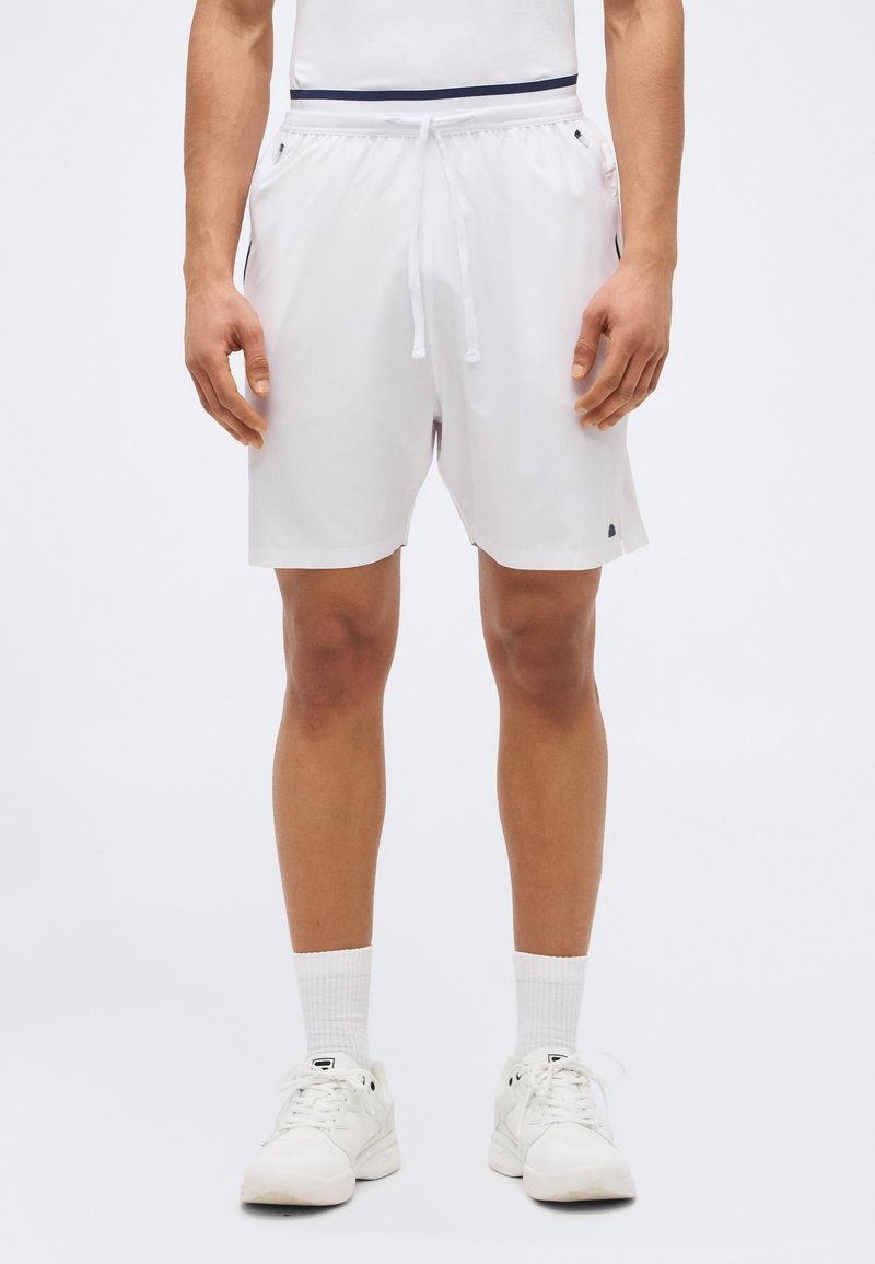 Άνδρας που φοράει λευκά αθλητικά σορτς με πλαϊνές τσέπες, λευκές κάλτσες κανονικού μήκους και λευκά παπούτσια, στέκεται μπροστά σε απλό φόντο.