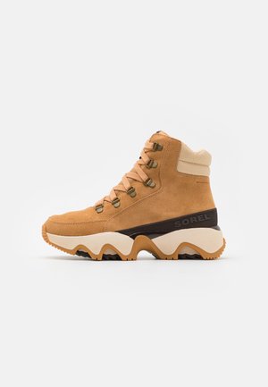 Tan-farbene Wildleder-Stiefelette mit beigen Akzenten, metallischen Ösen und einer klobigen weißen und braunen Sohle mit wellenförmigem Design. Mit "SOREL" gekennzeichnet.