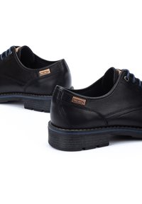 Chaussures en cuir noir avec des lacets bleus et une finition texturée. Présentent une étiquette de logo marron, des coutures contrastées et une semelle épaisse noire.