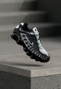 Μαύρο και άσπρο παπούτσι τρεξίματος Nike με μαλακή σόλα που αιωρείται πάνω από μια τσιμεντένια άκρη και σκάλες στο φόντο.