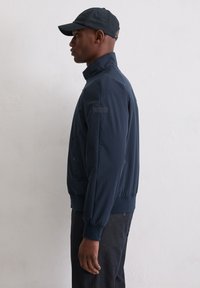 Granatowa kurtka bomber z wysokim kołnierzem, elastycznymi mankietami i bocznymi kieszeniami, zestawiona z dopasowaną czapką, na tle jasnej ściany.