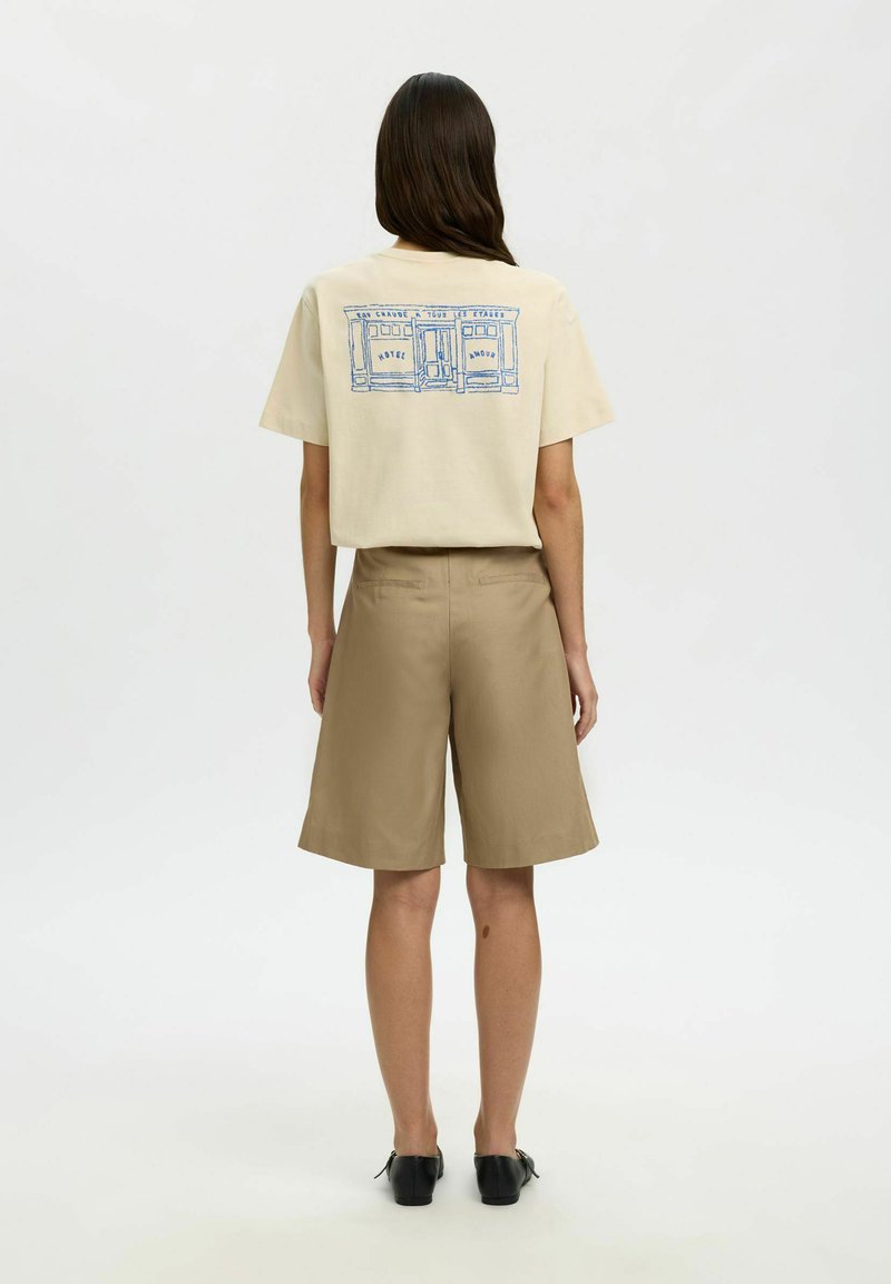 Femme aux longs cheveux noirs portant un short beige, des chaussures noires et un t-shirt crème avec un motif de devanture bleue au dos, debout de dos.