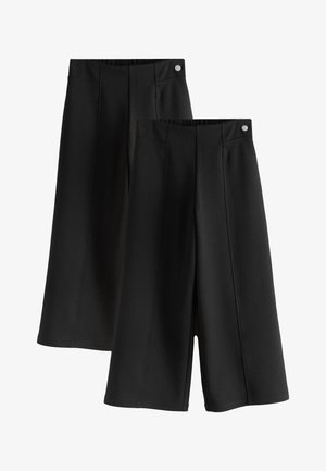 Mano sinistra che riposa accanto a pantaloni neri con cucitura e tasca, su sfondo bianco con parte di una camicia bianca visibile.
