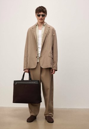 Noor mees, kes kannab beežist ülikonda ja päikeseprille, hoides suur mustast nahast ja kangast õlakotti ühtlase tausta ees.