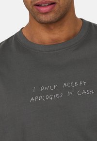 Gray cotton t-shirt with a round neckline. Features white embroidered text: "I ONLY ACCEPT APOLOGIES IN CASH." Smooth texture, casual design.