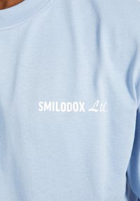 Camiseta de manga larga azul claro hecha de algodón; presenta el texto blanco "SMILODOX Ltd." en el pecho con una fuente simple y en negrita.
