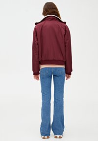 Burgunderfarvet isoleret jakke med høj krave, elastiske manchetter og talje, parret med flagrende blå jeans og hvide sneakers.