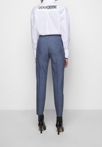 Pantalon bleu ajusté avec une coupe slim, présentant un tissage texturé et des poches arrière, assorti à une chemise blanche et des bottines à talons noires.