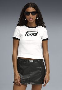 Biely skrátený tričko s čiernymi lemami a logom "PUMA", skombinované s čiernou koženou mini sukňou. Doplnky zahŕňajú malé čierne slnečné okuliare.