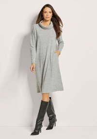 Graues Kleid mit Cowl-Neck und langen Ärmeln aus weichem Stoff, mit einem lockeren Schnitt und zwei seitlichen Taschen. Kombiniert mit knielangen schwarzen Stiefeln mit Absatz.