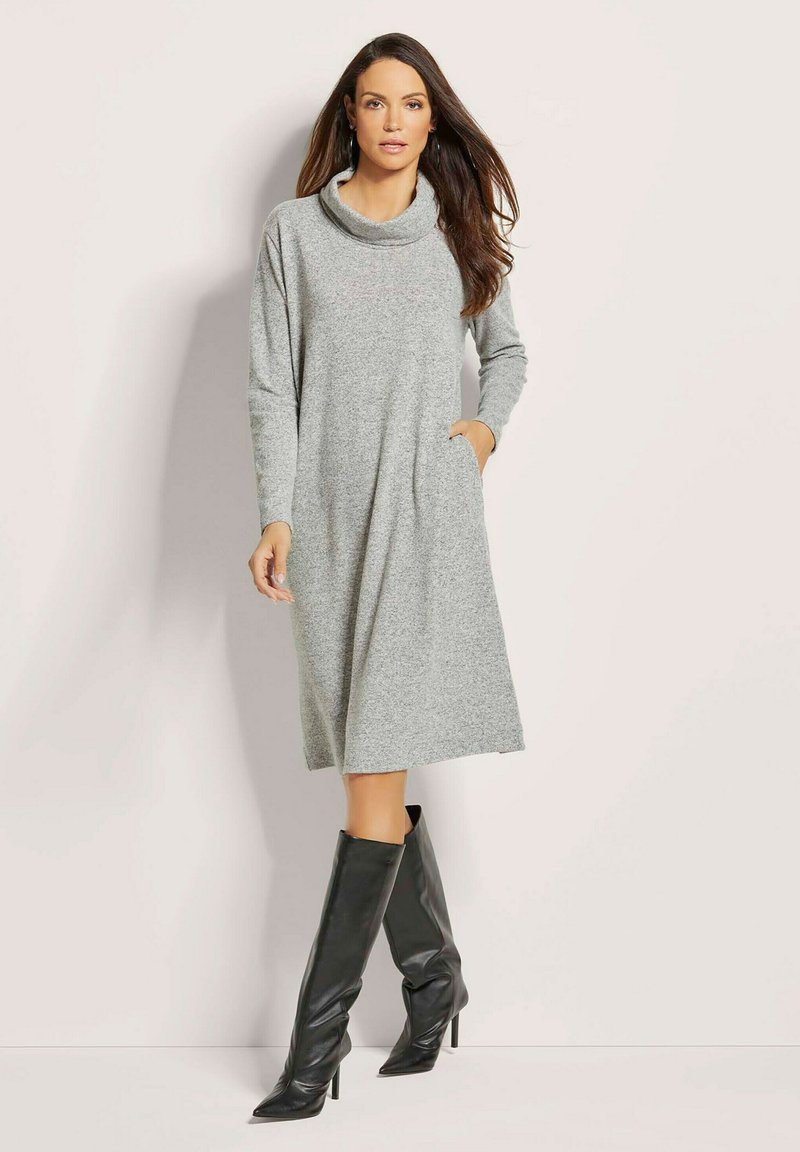 Graues Kleid mit Cowl-Neck und langen Ärmeln aus weichem Stoff, mit einem lockeren Schnitt und zwei seitlichen Taschen. Kombiniert mit knielangen schwarzen Stiefeln mit Absatz.