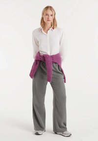 Mujer rubia de pie, vestida con una camisa blanca de botones, pantalones de pierna ancha a rayas grises, zapatillas blancas y un suéter morado atado a la cintura.