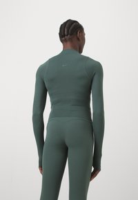Grünes sportliches Longsleeve-Oberteil mit figurbetontem Schnitt und flachen Nähten, mit einem dezenten Logo auf dem Rücken. Glatter, elastischer Stoff.