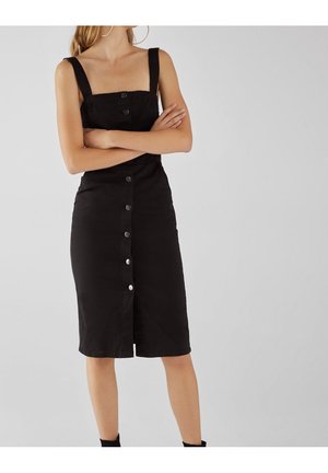 Vrouw met gekruiste armen die een mouwloze, zwart knielange jurk met knopen en brede banden draagt, tegen een effen grijze achtergrond.