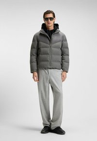 Grå puffjacka med en kontrasterande mörkgrå övre del, zip-öppning och justerbar huva; bärs över svart hoodie och ljusgrå byxor.