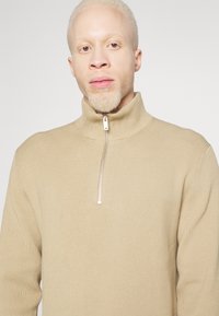 Maglione a coste beige con colletto alto e chiusura a zip metà. Texture morbida, vestibilità slim. Design minimalista senza ulteriori motivi o accenti.