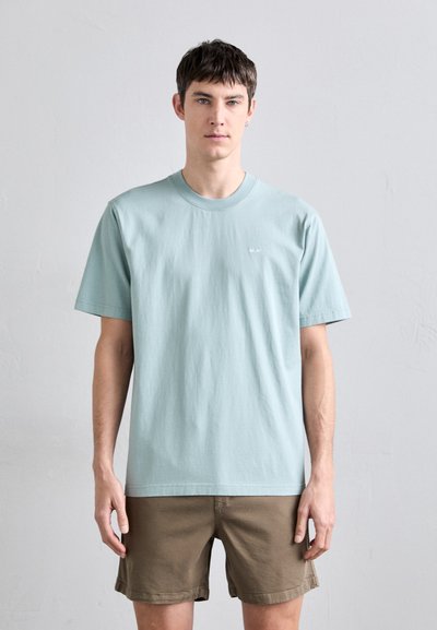 T-shirt in cotone azzurro chiaro con scollo rotondo e maniche corte. Presenta un piccolo logo sul petto. Indossato con pantaloni corti color khaki.