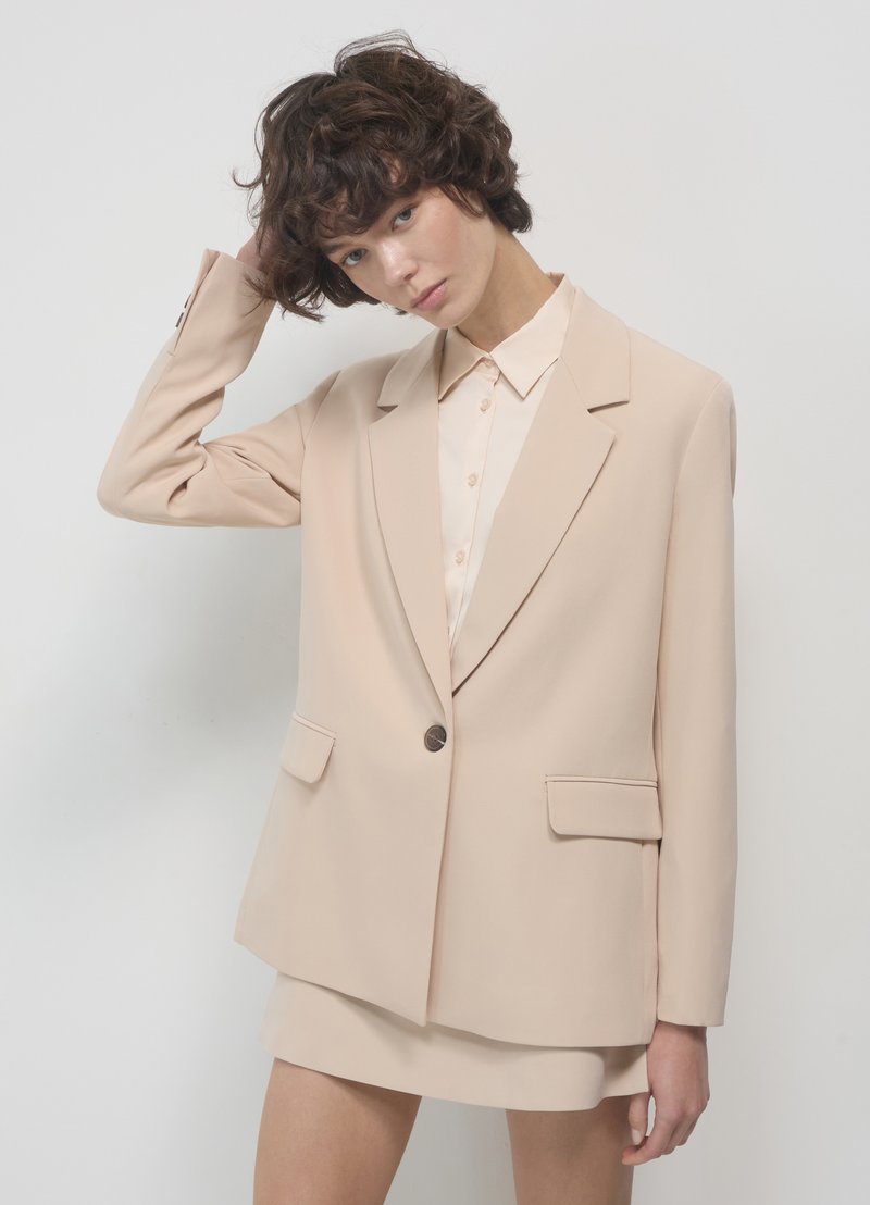 Blazer beige su misura con rever e due tasche frontali, abbinato a una blusa chiara. Texture liscia, design strutturato e dettaglio con un solo bottone.