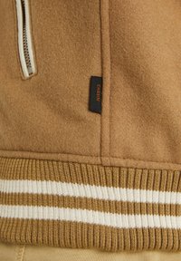 Brązowa kurtka polarowa z białym zamkiem błyskawicznym, czarną metką marki oraz ściągaczem z białymi i brązowymi paskami.