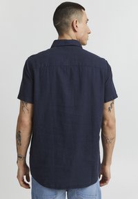 Camisa de manga corta de color azul marino, hecha de un tejido ligero, con cuello abotonado, dobladillo recto y un corte relajado. Tatuajes visibles en el brazo.