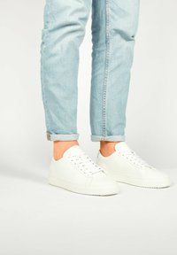 Baskets en cuir blanc avec un design minimaliste, bout rond, semelle plate, et six œillets avec des lacets, associées à un jean léger en bleu délavé.