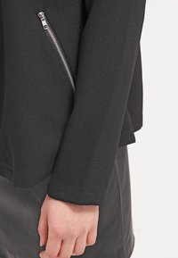 Veste noire en tissu texturé, coupe slim, avec une fermeture éclair argentée inclinée sur le côté gauche. Les extrémités des manches sont coupées droites.