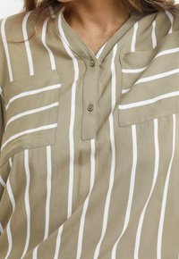 Chemise rayée beige et blanche à rayures verticales avec deux poches poitrine et un plastron à boutons partiel, portée par une personne.