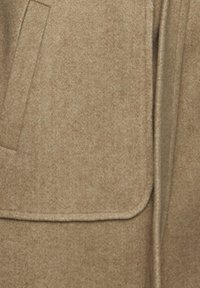 Bežani mješavina vune ima glatku teksturu s patch džepom i preklopom, naglašavajući minimalističke dizajnerske elemente.