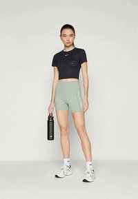 T-shirt sportivo nero accorciato abbinato a pantaloni corti fitted verde chiaro. Il modello tiene in mano una bottiglia d'acqua nera, indossando calze e scarpe bianche.