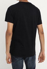 Černé bavlněné tričko s krátkými rukávy a kulatým výstřihem, pohled zezadu. Spodní okraj je mírně zakřivený. Nosené s modrými džíny.