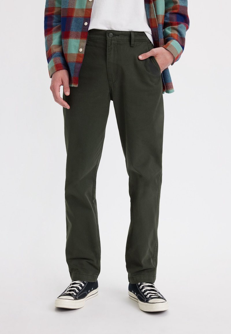Homme portant un pantalon droit vert foncé, une chemise à carreaux multicolores, un t-shirt blanc et des baskets en toile noires, la main dans la poche.