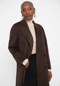 Barna gyapjú keverék, dupla soros kabát nagy hajtókákkal, fekete gombokkal és laza szabással. Bézs garbó fölött viselve.