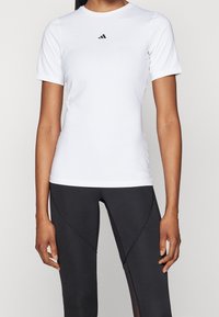 Kvinna iförd en figurnära vit t-shirt med kort ärm och svarta leggings med nätinsatser, som står mot en neutral bakgrund.