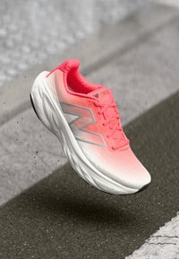 Une chaussure de course New Balance rose vif et blanche suspendue au-dessus d'une surface d'asphalte texture avec des marquages routiers blancs.