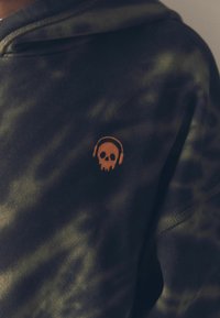 Schwarze und grüne Batik-Hoodie, mit einem orangefarbenen gestickten Schädel mit Kopfhörern. Weiche Textur, typische Kapuze und sichtbare Nähte.