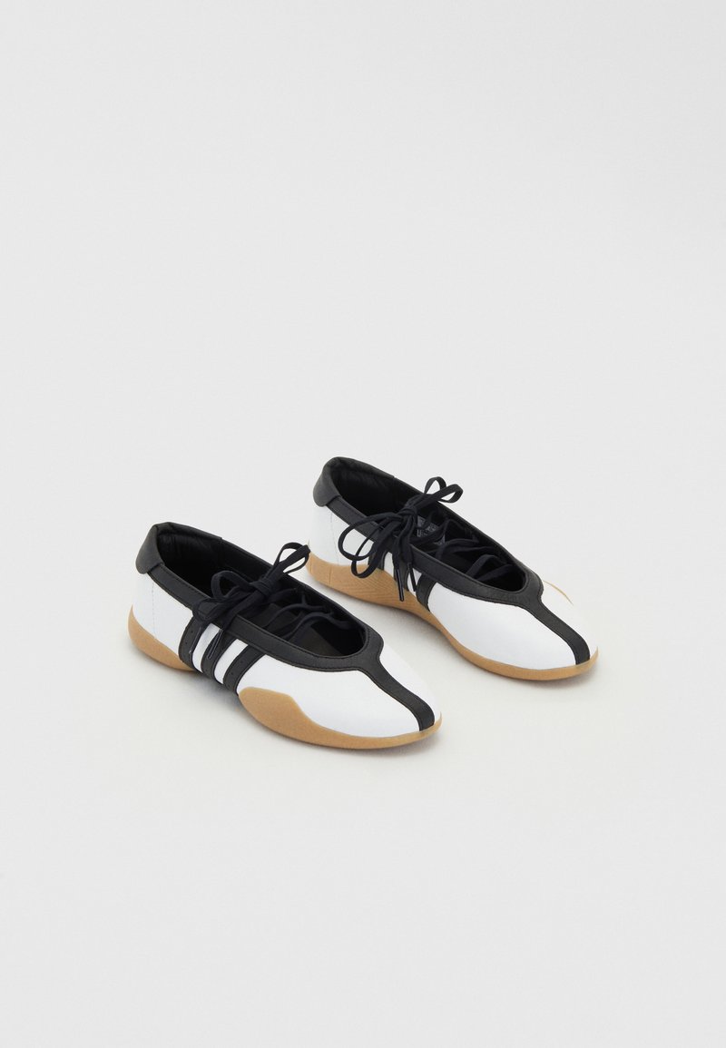 Sapatos brancos e pretos com atacadores e sola de borracha, apresentando um design elegante e três riscas pretas de lado. Textura suave.