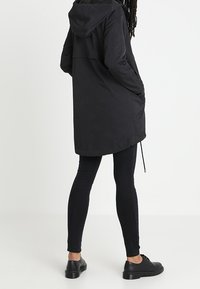 Personne portant un parka à capuche noir, un jean skinny noir et des chaussures à lacets noires, debout devant un fond blanc uni.