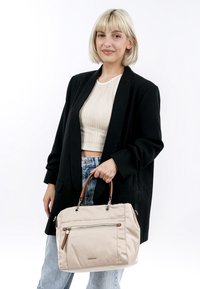 Beige tygväska med bruna läderdetaljer och silverfärgade metalldelar, med en framficka med dragkedja och dubbla handtag, bärs i en persons hand.