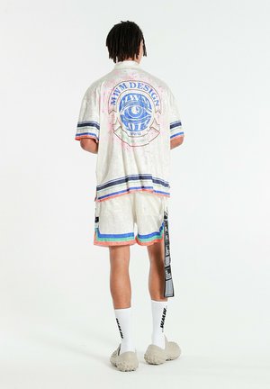 Hombre con rastas que lleva pantalones cortos blancos y camisa grande a juego con rayas azules y naranjas, diseño de ojo azul en la espalda y calcetines blancos.