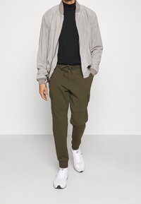 Ljust grå mockabomberjacka över en svart polotröja, kombinerad med olivgröna joggerbyxor med cargofickor och vita sneakers.