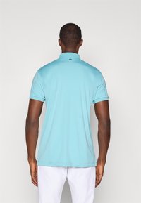 Lyseblå polo med ribbet krage, korte ermer, og logo-detalj på baksiden av nakken. Laget av et glatt, mykt stoff.